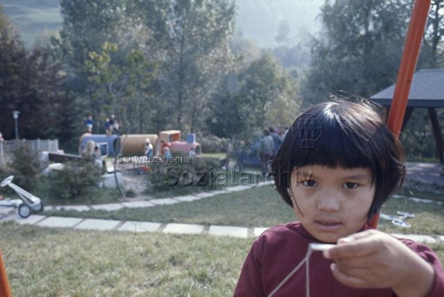 "Siedlungs- und Quartierspielplätze: Siedlungsspielplatz Lichtensteig SG" - Kleines Mädchen blickt in die Kamera, im Hintergrund sieht man spielende Kinder auf dem Spielplatz im Grünen; um 1970