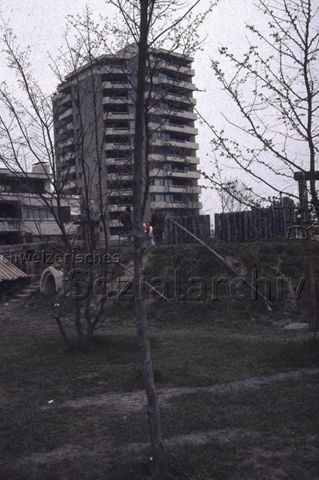 "Siedlungs- und Quartierspielplätze: Selbstbau Siedlungsspielplatz Moos, Oberhünenberg ZG, 1977" - Wiese mit jungen Bäumen und Spielgeräten, im Hintergrund ein hoher Wohnblock