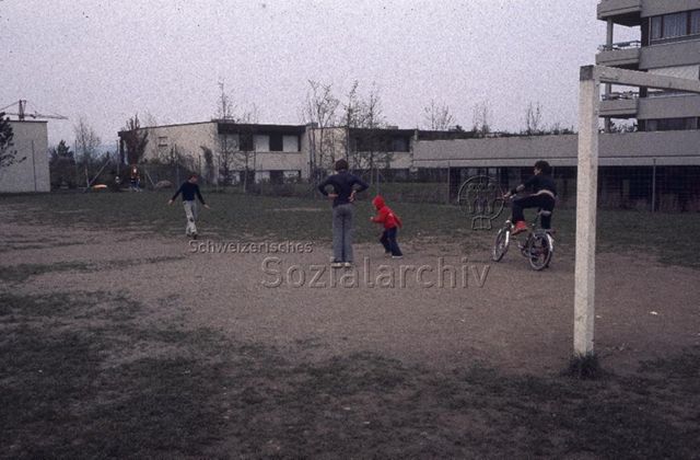 "Siedlungs- und Quartierspielplätze: Selbstbau Siedlungsspielplatz Moos, Oberhünenberg ZG, 1977" - Kinder spielen auf einem eingezäunten Rasenstück zwischen Wohnhäusern