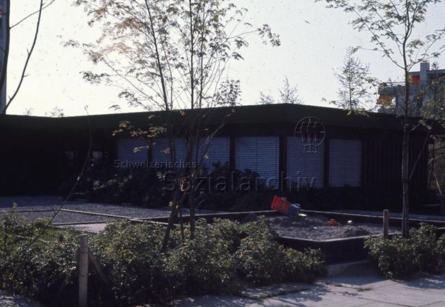 "Siedlungs- und Quartierspielplätze: Siedlungsspielplatz Hünenberg ZG" - Aussenansicht eines einstöckigen Gebäudes mit Sandkasten, Schottersteinplatz und Gartenstreifen; um 1970