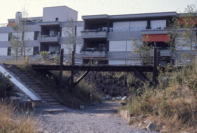 "Siedlungs- und Quartierspielplätze: Siedlungsspielplatz Hünenberg ZG" - Spielplatz mit Holzbrücke und Rutschbahn, im Hintergrund ein Wohnhaus; um 1970