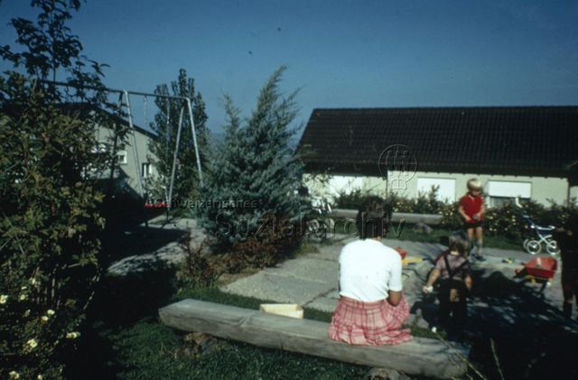 "Sitzplatz auf Siedlungsspielplatz" - Kinder unter der Aufsicht der Mutter am Spielen; um 1975