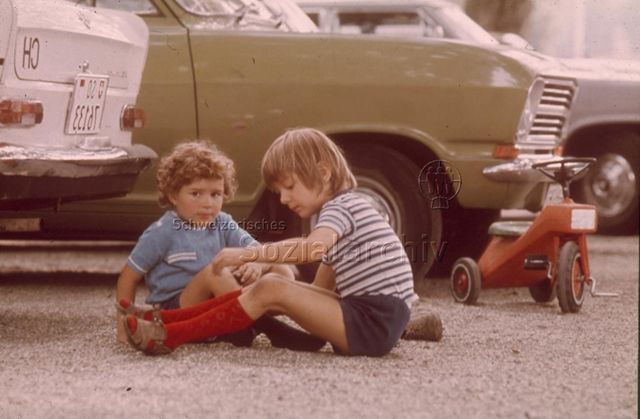 "Strasse als Spielplatz" - Zwei Kinder beim Spielen hinter geparkten Autos; um 1970