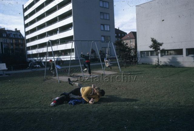 "Spielwiese" - Zwei Jungen beim Raufen, hinten wird geschaukelt; um 1975