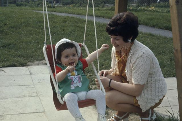 "Schaukel für Kleinkind" - Kleinkind sitzt in der Schaukel, die Mutter daneben; um 1975