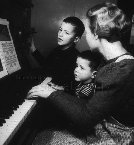 "Mutter singt mit ihren Kindern" und begleitet auf dem Klavier; um 1965
