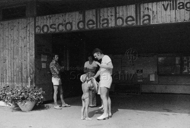 Bosco della Bella, Feriendorf für kinderreiche Familien - Ein Kind im Badeanzug und ein Mann mit Zigarette unterhalten sich, zwei Männer und ein Kind stehen daneben; um 1975