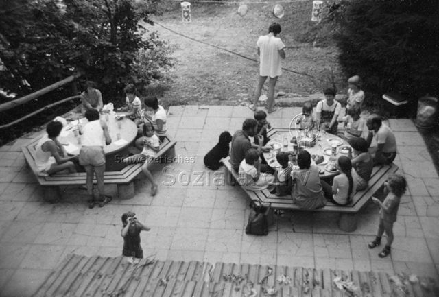 Bosco della Bella, Feriendorf für kinderreiche Familien - Eltern und Kinder sitzen um zwei Tische und essen und trinken; um 1975