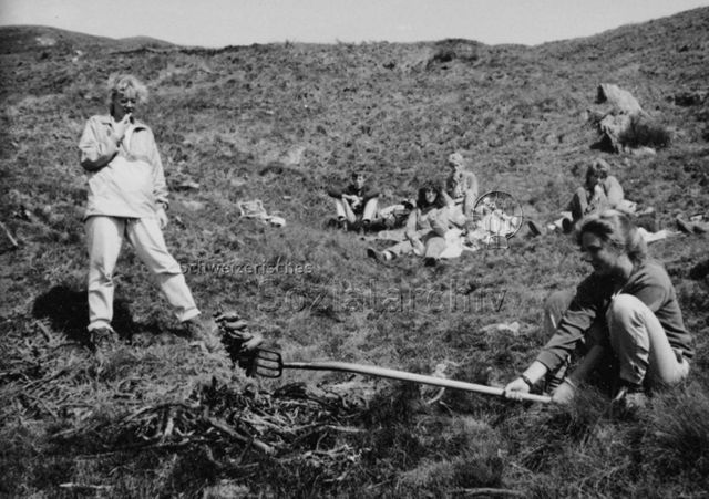 "Aktion 7, Tessin" - Jugendliche beim Ausruhen, vorne grillt eine Jugendliche Cervelats mit einer Forke; 1988
