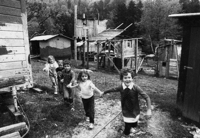 Spielplatz, Luzern - Kinder laufen händchenhaltend durch die Anlage, im Hintergrund die gebauten Hütten; um 1980