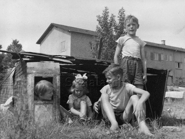 "Eine kleine selbstgebaute Hütte auf dem Bauspielplatz, die in der Phantasie der Kinder tausendmal schöner und heimeliger ist als das Wohnhaus im Hintergrund" - Vier Kinder in einer Hütte; um 1960