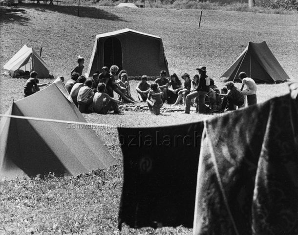 "Abenteuerlager Oberstammheim" - Jugendliche und Kinder sitzen um eine Feuerstelle zwischen den Zelten; um 1980