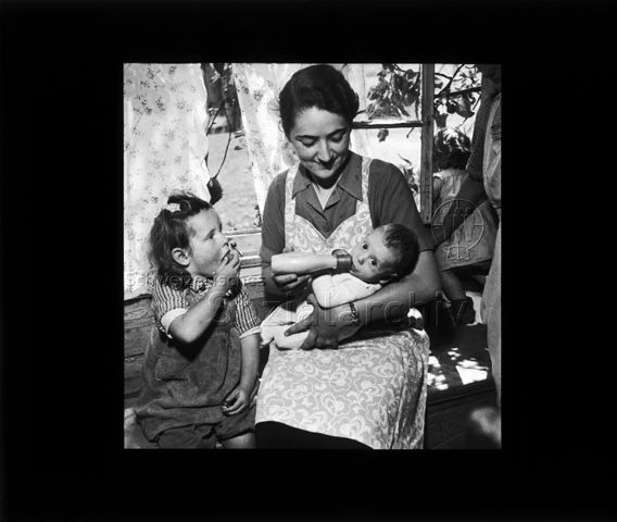 Diaserie "Reserve" und "alte Dias"; eine Frau und ein Mädchen sitzen auf einer Holzbank; die Frau mit einem Säugling im Arm, die Milchflasche gebend; das Mädchen mit Blick auf die Frau am Essen; um 1950