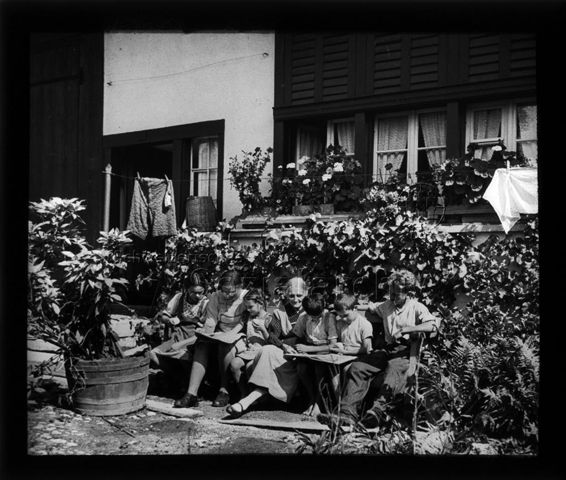 Diaserie zum Thema Mädchen; drei Mädchen und drei Jungen sitzen auf einer Sitzbank vor einem Haus, in der Mitte eine ältere Frau; um 1940
