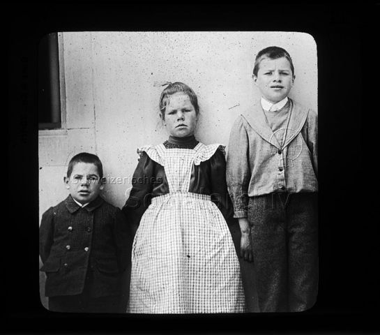 Diaserie zum Thema Kropfprophylaxe; "Kretinische Aargauer Kinder" - zwei Jungen und ein Mädchen mit Symptomen eines Jodmangelsyndroms stehen an einer Wand; um 1920