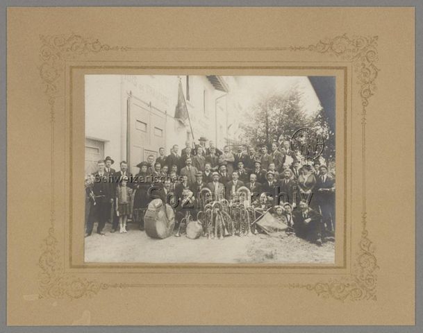 Musikgruppe (vermutlich Blasmusik von Viège), Fahnen, Blasinstrumente, Pauke, Trommel, Haus mit Beschriftung "Bois de chauffage, Moulin agricole", Fahne (rechts unten): "Concours de Martigny, Juin 1921, Musique de Viège"